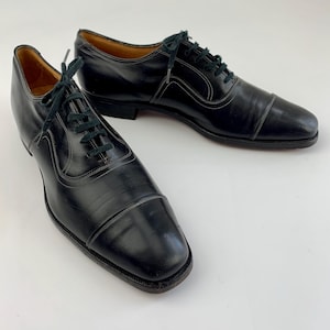 May include: A pair of black leather dress shoes with a cap toe design. The shoes have dark green laces and white stitching accents. The soles are dark brown. The shoes are displayed on a white background.