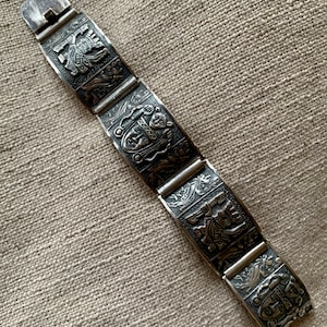 May include: A silver bracelet with detailed engraved designs. The bracelet is made up of rectangular links, each with relief carvings of figures and patterns. The clasp is a simple design. The bracelet is displayed on a textured, neutral-toned surface.