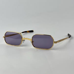 May include: Gold-framed sunglasses with rectangular purple lenses. The frames have a decorative design on the temples and a tortoiseshell pattern on the ear pieces. The sunglasses are set against a white background.