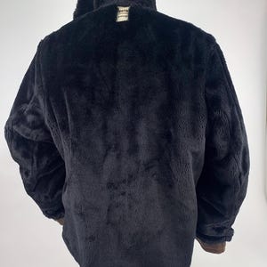 May include: Black faux fur coat with a large collar. The coat has a plush texture and a brown trim on the cuffs. A small tag is visible near the collar. The coat is displayed on a mannequin.