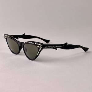 May include: Black cat-eye sunglasses with decorative embellishments along the top and sides of the frames. The sunglasses have a classic retro design with dark lenses and a sleek, modern look.