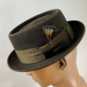 May include: A dark olive green fedora hat with a matching band and a decorative feather. The hat has a classic, structured shape with a slightly curved brim. The band is a lighter shade of olive green.