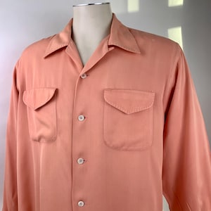 Puede incluir: Una camisa de manga larga color melocotón con dos bolsillos delanteros y cuello de campamento. La camisa tiene botones blancos en la parte delantera y en los puños. La tela parece ser un material suave y ligero.