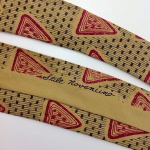 May include: A tan silk tie with a geometric pattern of red triangles and black lines. The tie has the text "Stile Noventimo" printed on it.