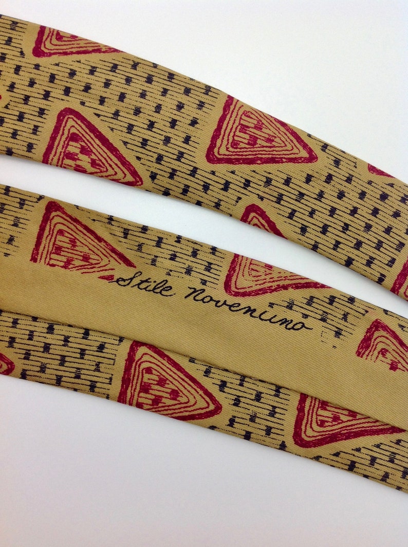 May include: A tan silk tie with a geometric pattern of red triangles and black lines. The tie has the text "Stile Noventimo" printed on it.