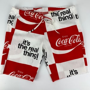May include: Red and white checkered shorts with the Coca-Cola logo and the text "Enjoy Coca-Cola, it's the real thing!" repeated throughout.