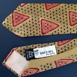 May include: A gold tie with a red and black geometric pattern. The tie has a label that reads "Oak Hall Memphis" and "All Silk RN 19881".