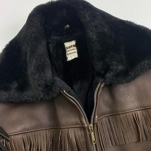 May include: Brown leather jacket with black faux fur collar and lining. Features a zipper closure and fringe detailing along the bottom. The label inside the jacket reads "UBER Owatonna Minnesota."
