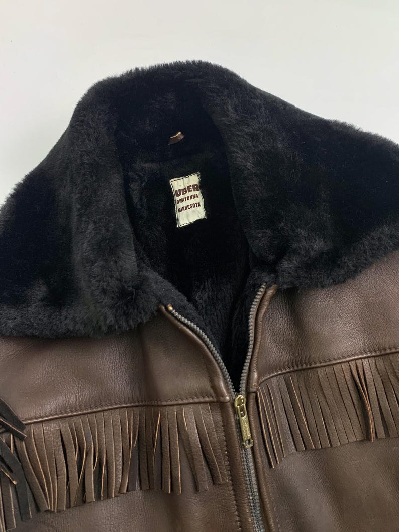 May include: Brown leather jacket with black faux fur collar and lining. Features a zipper closure and fringe detailing along the bottom. The label inside the jacket reads "UBER Owatonna Minnesota."