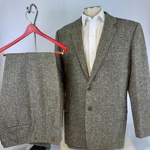May include: A two-piece suit in a grey tweed fabric with subtle flecks of red and blue. The blazer features a notched lapel, two front buttons, and a white collared shirt underneath. The matching trousers are displayed on a red hanger.