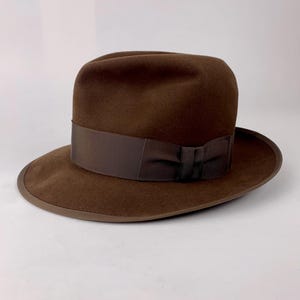 May include: A dark brown fedora hat featuring a structured crown with a centre crease and a pinched front. The hat includes a matching dark brown grosgrain ribbon band around the base of the crown and a finished edge along the brim.