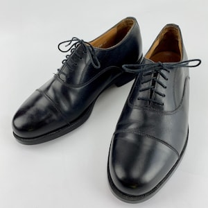 May include: A pair of black leather Oxford shoes with a classic cap-toe design. The shoes have a lace-up closure and a polished finish. The soles are dark, and the shoes appear unworn, set against a white background.