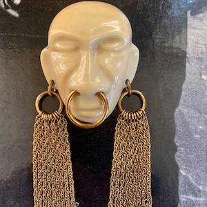 May include: A white ceramic face with a large gold ring through the nose and two gold hoop earrings with dangling chains. The face has closed eyes and a neutral expression.