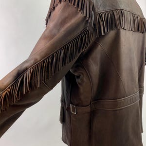 May include: Brown leather jacket with fringe detailing on the sleeves and back. The jacket has a black shearling collar, a belt at the waist, and a classic western style. The leather appears worn, giving it a vintage look.