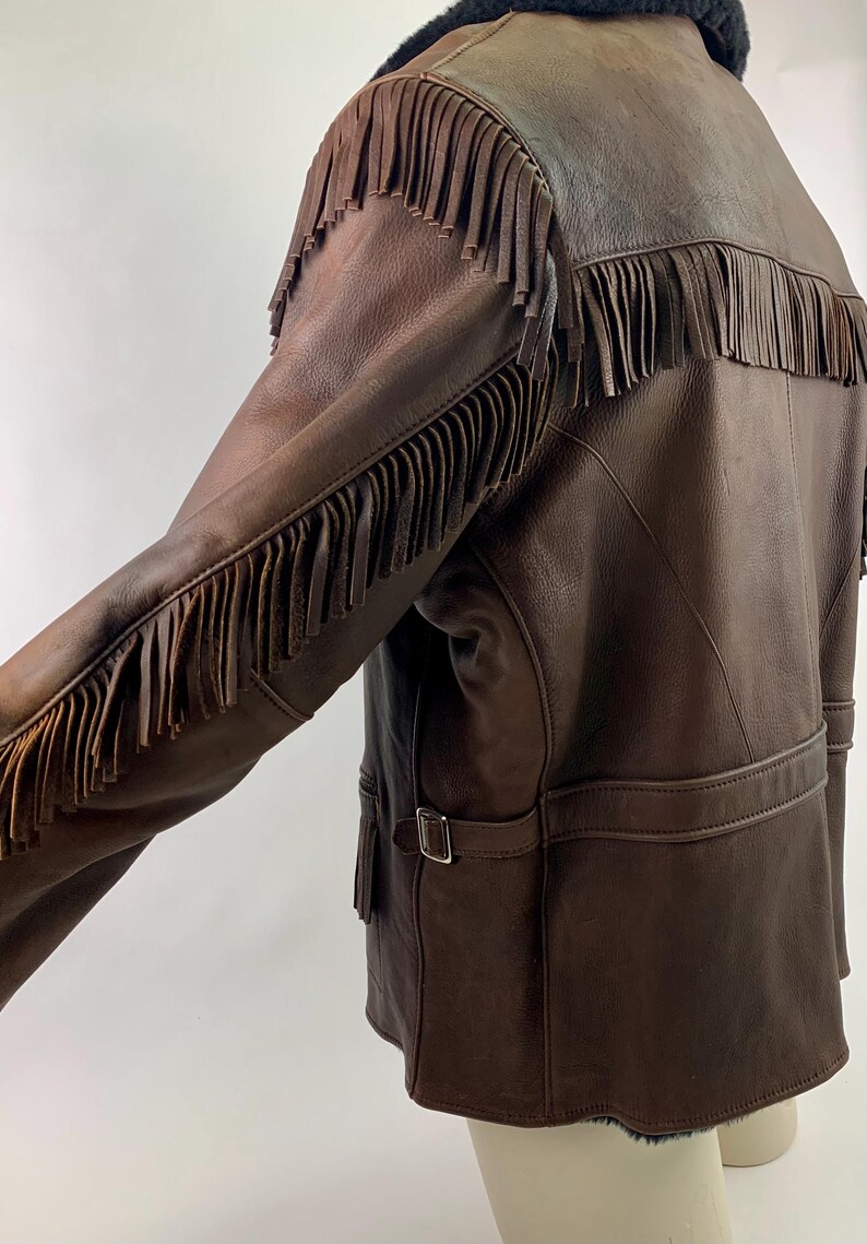 May include: Brown leather jacket with fringe detailing on the sleeves and back. The jacket has a black shearling collar, a belt at the waist, and a classic western style. The leather appears worn, giving it a vintage look.