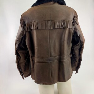 May include: Brown leather jacket with fringe detailing on the sleeves and back. The jacket features a black faux fur collar and a belt detail at the waist. The jacket is displayed on a mannequin.