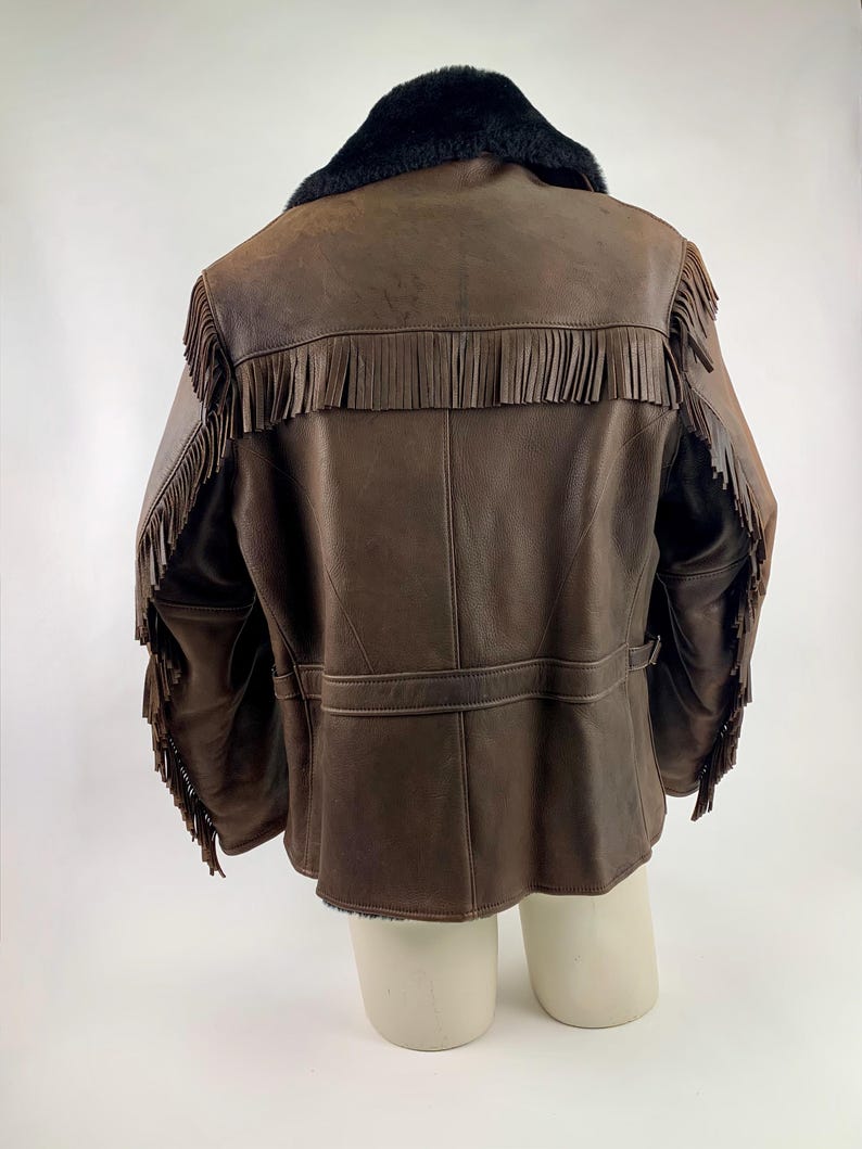 May include: Brown leather jacket with fringe detailing on the sleeves and back. The jacket features a black faux fur collar and a belt detail at the waist. The jacket is displayed on a mannequin.