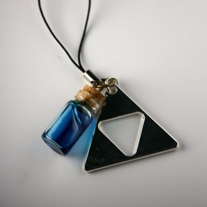 Könnte beinhalten: Eine blaue Glasflasche mit einem Korkenverschluss, der an einem silbernen Dreiecksanhänger mit einem kleineren ausgeschnittenen Dreieck befestigt ist. Der Anhänger ist an einer schwarzen Schnur befestigt.