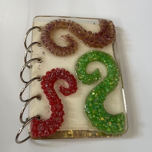 Op de afbeelding: Een klein, rechthoekig kunstwerk van hars met een spiraalbinding. Het stuk toont drie glitterende, gestileerde letter "S" vormen in rood, groen en bruin, tegen een crèmekleurige achtergrond.