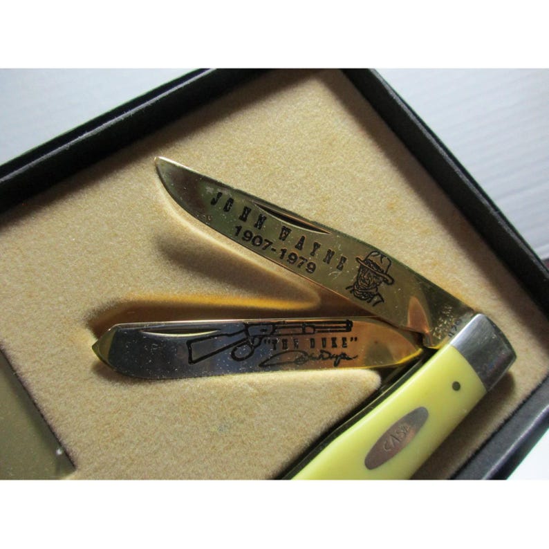 May include: A folding pocket knife with a yellow handle and two gold-toned blades. The top blade features the text "JOHN WAYNE 1907-1979" and a cowboy hat illustration. The bottom blade displays a rifle graphic, the text "THE DUKE", and a signature.