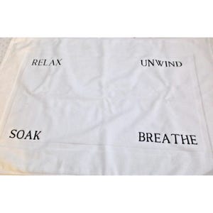 May include: White rectangular bath mat with the words "RELAX", "UNWIND", "SOAK", and "BREATHE" embroidered in black. The mat is made of a soft, absorbent material, perfect for a spa-like bathroom.