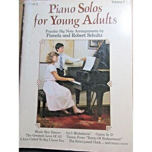 Peut inclure: Une couverture de livre avec le titre "Piano Solos for Young Adults" et le sous-titre "Popular Big Note Arrangements by Pamela and Robert Schultz". La couverture présente une jeune fille jouant d'un piano à queue noir avec un jeune garçon debout derrière elle.