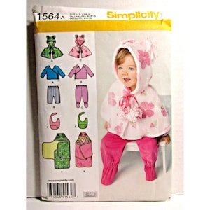 May include: Simplicity sewing pattern 1564A for infant clothing in sizes US XXS-L. The envelope features illustrations of a hooded cape, wrap top, pants, bib, and swaddle blanket, with a photo of a child wearing a white hooded cape with pink flowers.