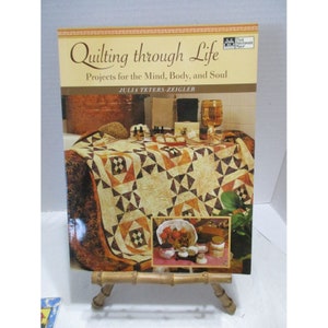Puede incluir: Un libro titulado "Quilting through Life: Projects for the Mind, Body, and Soul" de Julia Teters-Zeigler. La portada presenta una colcha con un patrón marrón, naranja y blanco, y una foto de un baño con una bañera de cobre y una copa de vino.