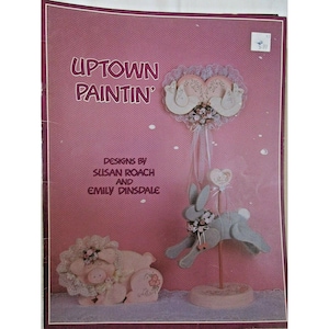 Puede incluir: Una portada de libro de manualidades rosa con el título "Uptown Paintin'" y los nombres Susan Roach y Emily Dinsdale. La portada presenta diseños decorativos de animales de fieltro, incluyendo un cerdo, un conejo y un diseño en forma de corazón.