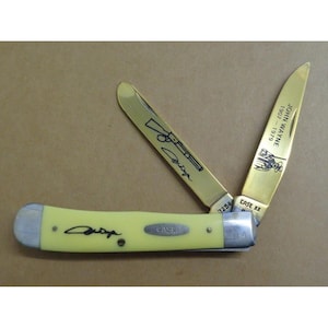 May include: A folding pocket knife with a pale yellow handle featuring a black signature. Two gold-toned blades are extended; one displays a rifle illustration and signature, while the other shows the text JOHN WAYNE 1907-1979 and a figure.