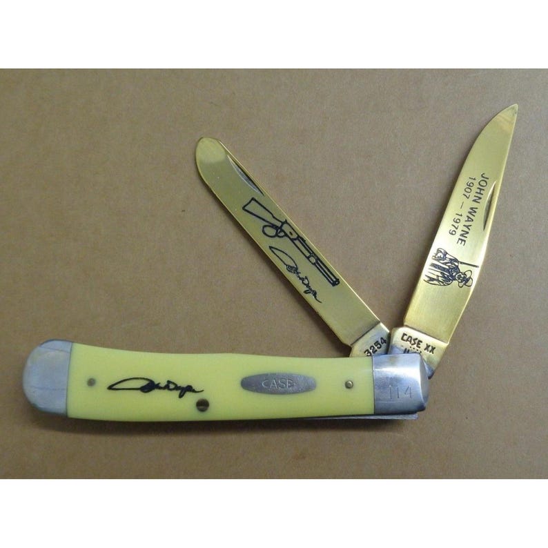 May include: A folding pocket knife with a pale yellow handle featuring a black signature. Two gold-toned blades are extended; one displays a rifle illustration and signature, while the other shows the text JOHN WAYNE 1907-1979 and a figure.