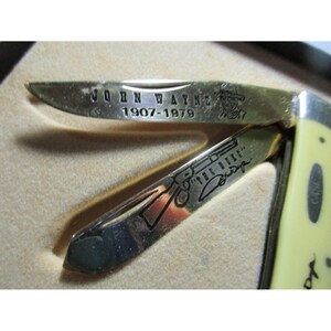 May include: A folding pocket knife with two metallic blades featuring engravings. The top blade displays the text JOHN WAYNE 1907-1979 and a small portrait. The bottom blade shows a rifle illustration, the text THE DUKE, and a signature. The handle is cream.