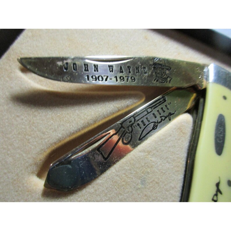 May include: A folding pocket knife with two metallic blades featuring engravings. The top blade displays the text JOHN WAYNE 1907-1979 and a small portrait. The bottom blade shows a rifle illustration, the text THE DUKE, and a signature. The handle is cream.