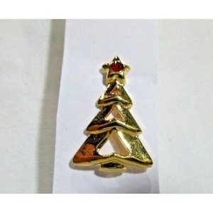 May include: A gold-coloured Christmas tree brooch with a red star on top. The brooch has a layered design, with each layer of the tree shaped like a triangle. The brooch is likely made of metal and is set against a white background.