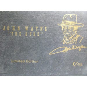 May include: A dark gray rectangular box featuring gold text that reads JOHN WAYNE THE DUKE, a gold line drawing of John Wayne wearing a cowboy hat, a gold signature, the text Limited Edition, and a small gold Case logo in the bottom corner.