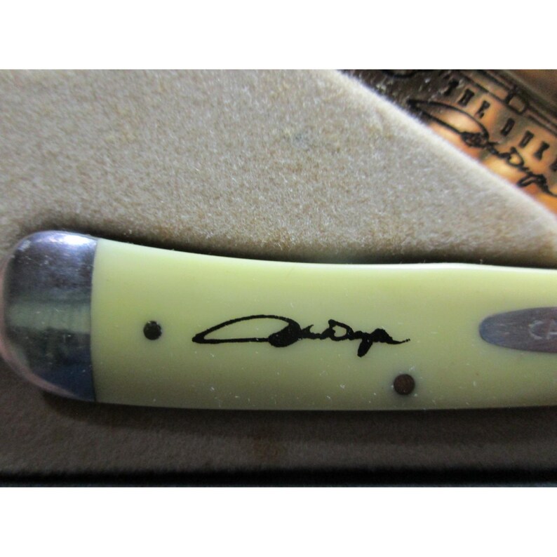 May include: A pale yellow pocket knife handle featuring a black signature printed on the surface. The handle has a metallic bolster on the left side and two small dark pins visible. A portion of a metallic blade is visible in the upper right corner.