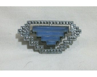 May include: Vintage Art Deco brooch with a geometric design. It features a light blue central element, framed by a silver-toned metal setting with small, sparkling accents. The brooch is an elongated oval shape.