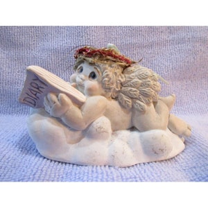 May include: A white cherub figurine with wings, wearing a red and brown wreath, lies on a cloud and holds a purple book that says "DIARY" on the cover.
