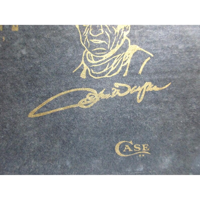 May include: A dark gray textured surface featuring a gold-colored line art illustration of a person wearing a scarf, a gold-colored signature that reads John Wayne, and a gold-colored logo that reads CASE with two small X symbols underneath.