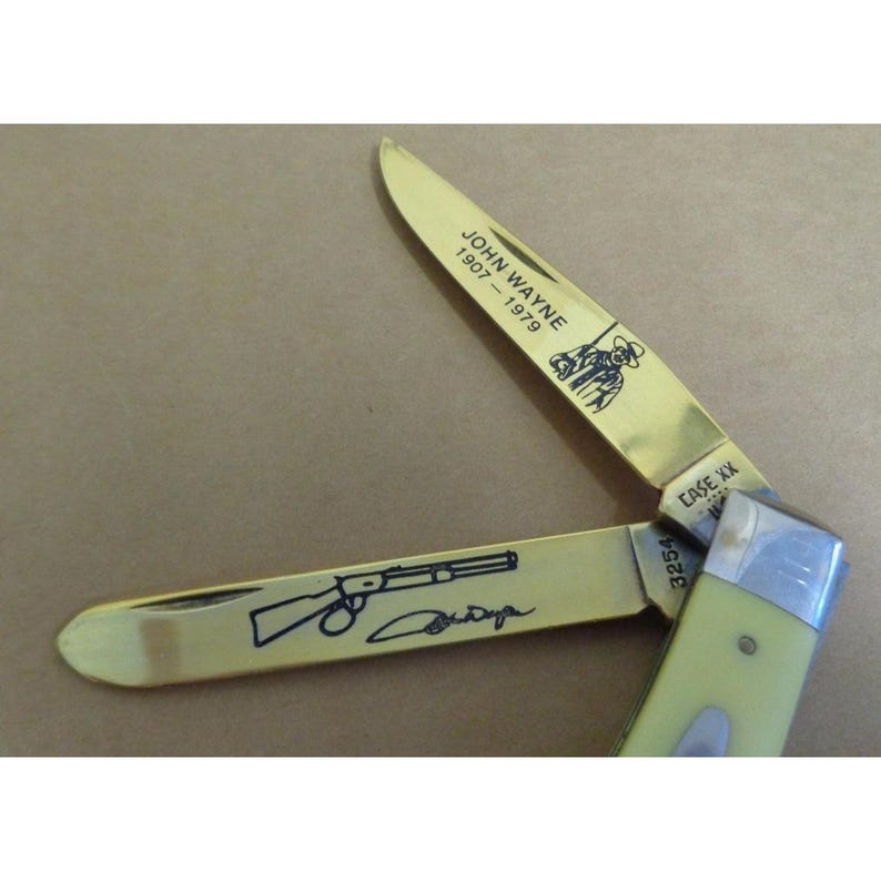 May include: A folding pocket knife with two brass-colored blades and a cream-colored handle. One blade features the text JOHN WAYNE 1907-1979 and an illustration of a person. The other blade displays a rifle illustration, a signature, and the number 3254.