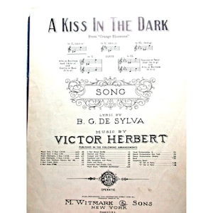 Puede incluir: Partitura de la canción "A Kiss in the Dark" de "Orange Blossoms". La portada presenta notación musical, la palabra "SONG" en un marco decorativo y los nombres B.G. De Sylva y Victor Herbert. Publicado por M. Witmark & Sons, Nueva York.
