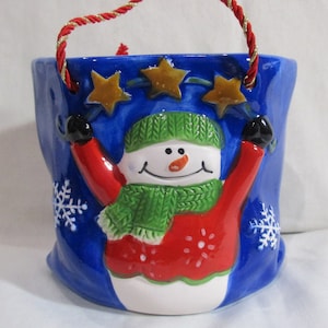 Op de afbeelding: Een blauwe keramische sneeuwman hangende plantenpot met een rood-groene sjaal en een rode muts. De sneeuwman houdt twee gouden sterren omhoog. De pot heeft een rood en gouden touwhandvat.