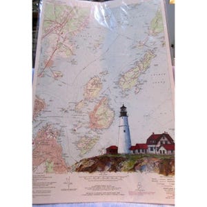 May include: A vintage map of Portland, Maine, featuring a detailed depiction of the coastline, islands, and surrounding waters. The map includes a lighthouse illustration with a white tower and red roof, set on a rocky cliff. The map is protected by a clear plastic sheet.