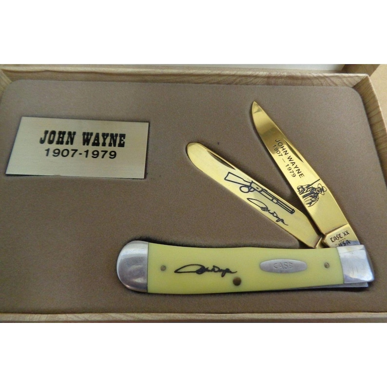 May include: A folding pocket knife with a pale yellow handle and two gold-toned blades. One blade features an engraving of a rifle and John Wayne's signature. A plaque in the box reads JOHN WAYNE 1907-1979. The knife is set in a tan display case.