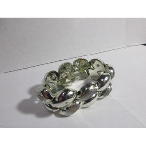 May include: Silver-toned stretch bracelet featuring a repeating pattern of linked, stylized seashell shapes. The bracelet has a polished, reflective surface and is designed to expand and contract for easy wear. A classic piece of jewelry.