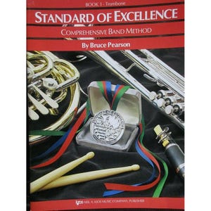 May include: A book titled "STANDARD OF EXCELLENCE" with the subtitle "COMPREHENSIVE BAND METHOD" by Bruce Pearson. The image includes a trombone, a trumpet, a medal in a box, and drumsticks. The medal has red, green, and blue ribbons.