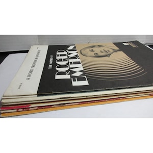 Pode incluir: Uma pilha de álbuns de discos de vinil vintage. A capa do álbum superior é preta com o texto "The Music of Roger Emerson" e um retrato. Outra capa do álbum diz "A Word From Our Sponsor".