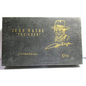 May include: A rectangular black box featuring gold text and graphics. The text reads JOHN WAYNE THE DUKE, Limited Edition, and CASE. A gold line drawing of John Wayne wearing a cowboy hat is present, along with a gold signature below the portrait.