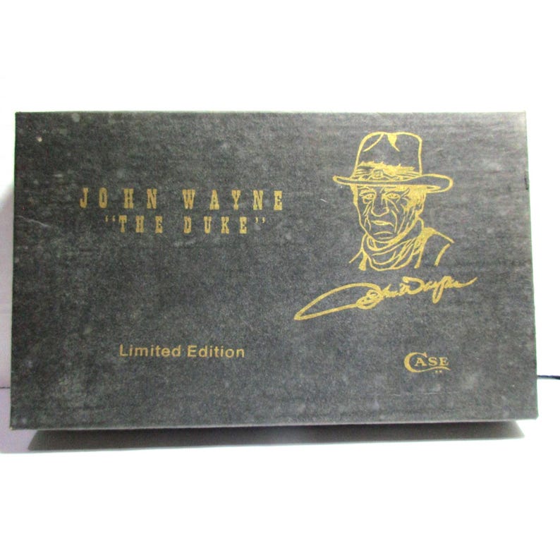 May include: A rectangular black box featuring gold text and graphics. The text reads JOHN WAYNE THE DUKE, Limited Edition, and CASE. A gold line drawing of John Wayne wearing a cowboy hat is present, along with a gold signature below the portrait.