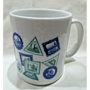 May include: White ceramic mug with a blue and green design of various stamps featuring different cities and states. The stamps feature images of buildings and landmarks.